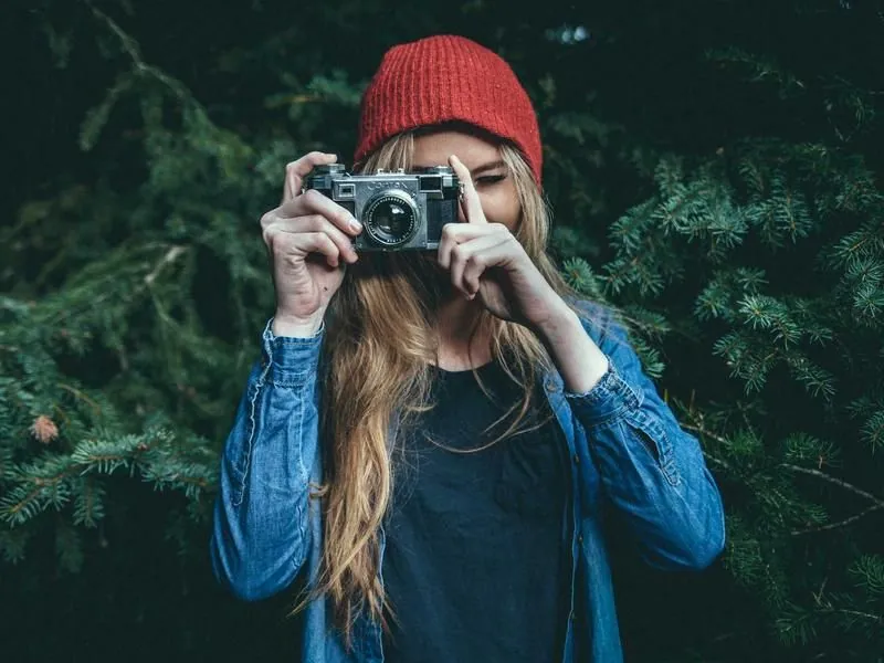 Original photo: a woman in a red beanie holding a camera in front of dark green foliage