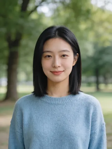 A portrait photo of a young woman with shoulder-length black hair and a light blue sweater, standing in a park