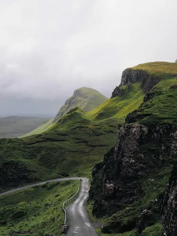 A photo of a winding road between green hills and dark rock faces, under a heavy overcast sky